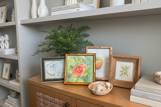 Framed artworks including rose original watercolor on a cabinet with a plant in the background
