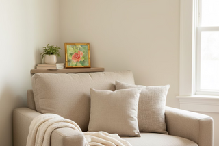 Beige armchair with cushions and a small table with a plant and framed picture in a room with a window.