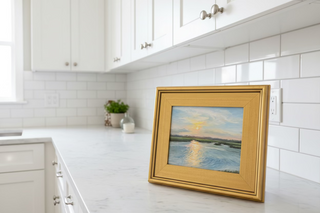 Gold-framed painting of a sea/marsh landscape with a lamp and plant in the background
